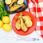 An image showing lemon pepper chicken drumsticks on a red plate with a red buffalo check napkin and lemons