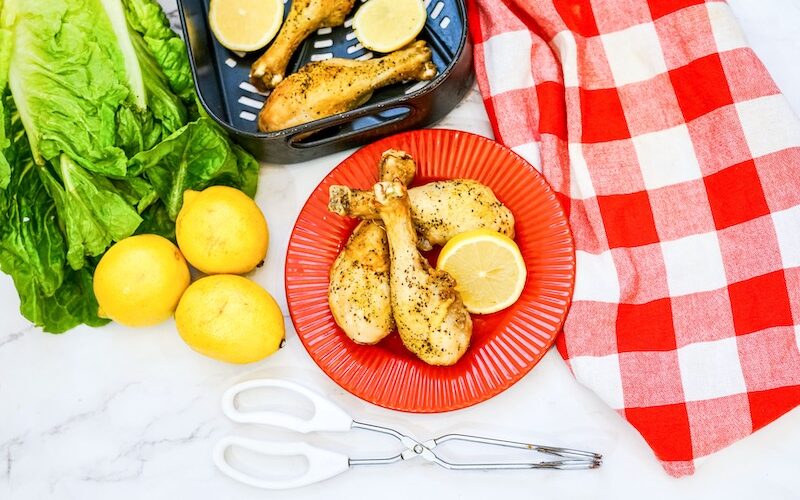 An image showing lemon pepper chicken drumsticks on a red plate with a red buffalo check napkin and lemons